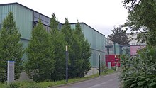 Street leading to the red entrance of the Max Planck Institute for Gravitational Physics in Hannover. The building is partly hidden behind trees next to the street.