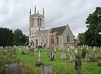 The tower of a stone church, seen from the west, at the top of which is a battlemented parapet with pinnacles. In front of the church and to the left is a lychgate