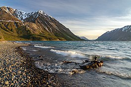 Small waves breaking on a rocky beach of a large body of water, with golden late-evening sunlight on snow-capped peaks in the distance