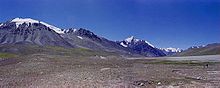 Some tall, bluegrey mountains rise out of brown soil below a deep blue sky