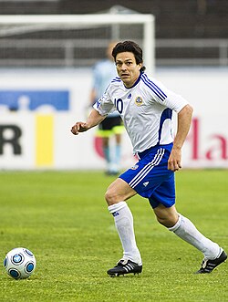 A photograph of a man on a football pitch wearing a white football shirt, blue shorts and white socks. The man is in possession of the football and glancing forward for something or someone out of picture.