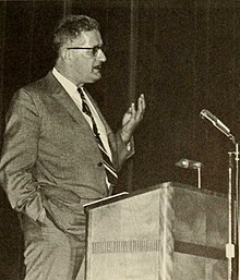 Theodore Ropp speaking at a podium and gesturing with his left hand