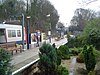 A grey railway platform with a series of light poles surrounded by brown trees, green bushes, and yellow and white flowers all under a white sky