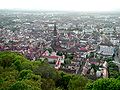 A view of Freiburg's old city taken from a hill. There are lush green trees in the foreground. In the middle ground, the Freiburg Minister is visible, as are older buildings. There are buildings out to the horizon, but the entire scene is very calm.