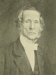 A black-and-white portrait of an elderly man with wavy side-parted hair, wearing a high-collared shirt with a cravat and a dark suit, gazing directly at the camera with a serious expression.