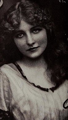 A young white woman, wearing a striped or pleated dress with a scoop neckline.