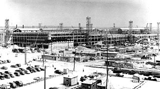 Expansive view of a construction site with lots of parked cars, scaffolding and cranes. There are a number of demountables in the foreground.