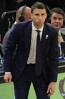 Full length view of man leaning forward, standing on side lines, wearing white shirt, dark tie and suit, scorers or announcers visible behind him