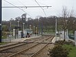 A set of two tram tracks between two platforms with shelters and a level pedestrian crossing between the two platforms. Just before the horizon there is a road which the tracks cross into.