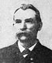 Head of a white man with a large drooping mustache and receding hairline, wearing a dark suit coat over a shirt and tie.