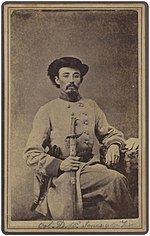 Sepia photo shows a seated man with a moustache wearing a black hat and a light gray military uniform.