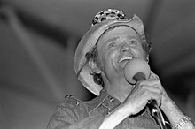 A black and white photograph of a smiling man wearing a cowboy hat and holding a microphone
