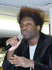 A Black man with tall hair is wearing a grey t-shirt and black coat; he is looking behind the camera, speaking into a microphone.