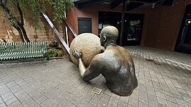 A rear view of the 'Inclined' sculpture, showing a muscular figure pushing a large, textured sphere. The figure's detailed back and arm muscles are prominent as they exert effort, emphasizing the theme of perseverance and struggle.