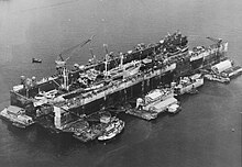 A floating drydock containing ships. It is surrounded by floating barges with workshops and a tugboat.