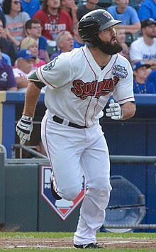 A man wearing a black batting helmet and white baseball uniform with "Sounds" written across the chest in red lettering runs on a baseball field after hitting the ball