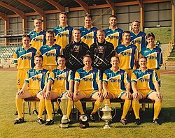 A group of young men in blue and yellow sports kit displaying several trophies