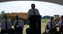 Transportation Secretary Ray LaHood stands at a podium flanked by Arkansas state officials and addresses those attending the groundbreaking with a large dump truck in the background