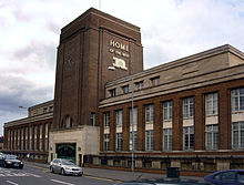 A long three-storey building made of brown brick. It has large windows and its tall central square tower provides a fourth storey. The side of the tower facing the road has large decorative iron gates at street level, and a simple clock closer to the top. The words "Home of the Best", above the stylised lowercase letter 'n' representing Nottinghamshire County Council, are on the other side of the tower that is visible.