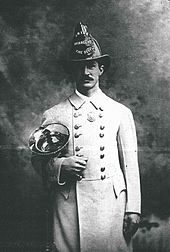 A man in a white coat and fire helmet holding a brass horn