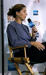 A brown haired woman, in black pants and blue shirt with a microphone in hand