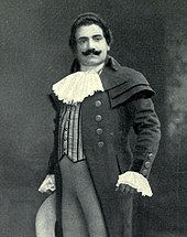 Man, with dark hair and a curling moustache, standing in a posed position. He is wearing a long coat, with lace at the throat and cuffs.