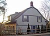 A gray wooden house seen from the side, lit by a setting sun from the left. Its roofline curves in segments, with a brick chimney at the crest. The upper section has wood shingled siding in a scale pattern.