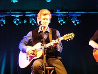 Singer Rodney Crowell sitting on a stool, singing into a microphone while strumming a guitar.