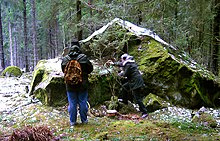 Two people with their backs to the viewer stand in front of a large boulder in the middle of woodland. One of the figures is pouring a liquid onto the ground.