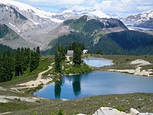 A forested, cone-shaped hill overshadowed by glaciated mountains rising over two pond-like lakes in the foreground.