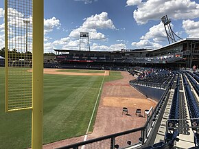 A green baseball field surrounded by its brown shale warning track and blue seats viewed from the concourse by the yellow left field foul pole on a sunny afternoon