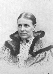 B&W portrait photo of a middle-aged woman, hair parted in the middle and pulled back, wearing a fancy coat.
