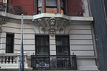 Detail of a balcony on the second story. The balcony has an ornate black-metal railing. Behind it is a window.