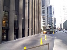 The recessed plaza surrounding the CBS Building, looking west along 53rd Street. The plaza has gray pavement and is several steps below the street. There are yellow cones in the plaza. To the left is the CBS Building, and to the right is the sidewalk on 53rd Street.