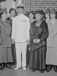 B&W image of Guglielmo Marconi and Edna Owen standing with Hunter college wireless students. Marconi is standing at attention and wearing a white uniform (shoes to cap). Owen dressed in a full-length dress with hat. Her hands are clasped in front of her holding a handbag.