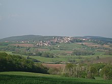 A small village with a visible church tower, on a low hill surrounded by fields and trees, seen from another such hill