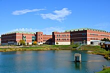 A large red brick multi-story building sits behind a lake on a mostly clear day.