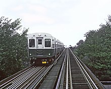 An unsaturated photograph of a train painted green on the bottom and white on the top, viewed head-on on the viewer's left track