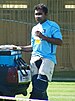 A man in a blue t-shirt, wearing white tracksuit trousers with pads. A grass-cutting machine can be seen in background.