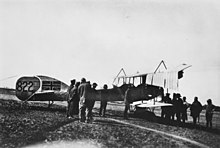 A group of men gather around a biplane. The Union Jack has been painted on to the tail of the aircraft.