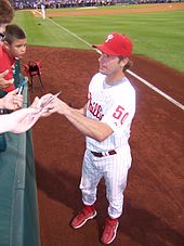 A man in a white baseball uniform with red lettering and a red hat and red shoes stands on a baseball field reaching into the stands to sign autographs for fans.