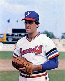 A man in a blue baseball cap with a red bill and a white "N" on the front and a white jersey with "Sounds" across the chest in red and blue stands on a field with his hands together in a brown leather glove.
