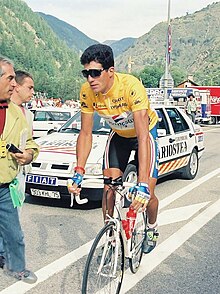 A man with dark hair stood up riding a bicycle wearing a yellow jersey