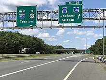 A four lane freeway at an interchange in a wooded area with two green signs over the road. The left one reads Interstate 195 west Trenton and the right one reads exit 21 County Route 527 County Route 526 Jackson Siloam upper right arrow