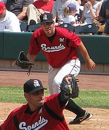 A man with a black cap, red baseball jersey with "Sounds" written across the chest in white, and white pants leans forward at first base preparing to catch a ball.