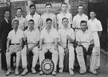 A group of 11 young cricketers in an official photo. They are wearing their white cricket uniforms, and five sit in the front row, and six stand behind them, along with a middle-aged man, their coach, in a dark suit and tie. Some are holding bats and wearing pads and a shield is bat the foot of the central boy in the front row.