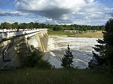 Dam wall shown with water flowing out three of the five floodgates. Spectators are gathered on the viewing area on the top of the wall watching the water flow past.