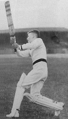 A male youth wearing cricket pads, gloves, white uniform and sweater, stands on the grass and follow through after swinging his bat in a vertical arc.