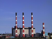 Photograph of four tall smokestacks with alternating red-white stripes, with a final black stripe at the top, in front of an industrial building. The fourth smokestack is half as tall and has no black stripe.