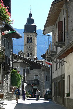 View in bright sunlight along a narrow street with stone buildings, pedestrians and church in background against a blue sky and a dark mountain in background